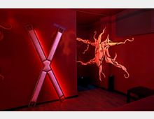Ein dunkelrot beleuchteter Raum mit rotem Holzboden und Wänden, darauf eine große, leuchtende X-förmige Struktur und eine abstrakte Dekoration in Flammenform mit leuchtenden Konturen. Im Hintergrund befindet sich ein dunkler Nischenbereich.