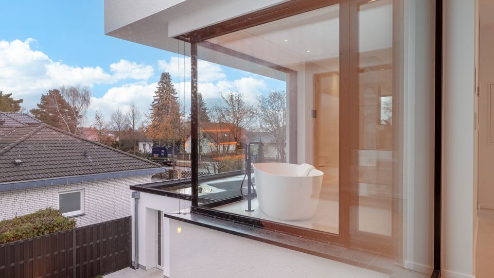 Moderne Wohnhausfassade mit bodentiefen Glasfronten und Holzrahmen, die einen hellen Innenraum mit freistehender Badewanne zeigen. Im Hintergrund sind ein Wohngebiet mit Ziegeldächern, Gartenzaun und Bäumen unter blauem Himmel zu sehen.