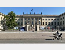Die Frontansicht der Humboldt-Universität in Berlin zeigt ein klassizistisches Gebäude mit Säulen am Eingangsbereich. Vor dem Gebäude sind ein schmiedeeisernes Tor, eine Statue und Informationsplakate, während Passanten und ein Fahrradfahrer die Straße davor überqueren.