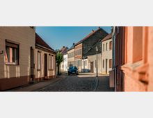 Schmale Kopfsteinpflasterstraße in einem Wohngebiet mit dicht stehenden, niedrigen Häusern mit Putz- und Ziegelfassaden sowie roten Satteldächern. Entlang der Straße stehen eine Laterne und ein geparktes Auto, im Hintergrund setzt sich die Häuserzeile fort.