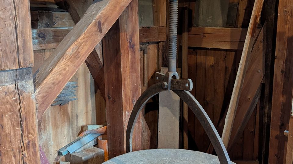 Innenraum einer historischen Mühle mit hölzerner Balkenkonstruktion und grob vertäfelten Wänden. Im Vordergrund steht ein großer runder Mühlstein mit zentraler Öffnung und Metallbügel, daneben sind alte Zahnräder und mechanische Bauteile sichtbar.