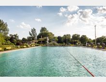 Freibad mit großem, rechteckigem Schwimmbecken, umgeben von Bänken, Strandkörben und Bäumen im Hintergrund; eine Rutsche mit Wendeltreppe ist auf der rechten Seite sichtbar. Der Himmel ist leicht bewölkt, und die Umgebung wirkt grün und parkähnlich.