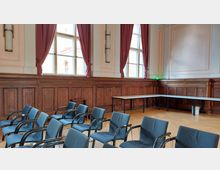 Ein heller Konferenzraum mit hohen Fenstern, roten Vorhängen und holzvertäfelten Wänden. Im Vordergrund stehen blaue Stühle in Reihen, im Hintergrund sind weiße Tische auf hellem Parkettboden zu sehen.