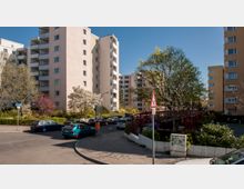 Wohngebiet mit mehrstöckigen Wohnhäusern aus hellen Fassaden, umgeben von blühenden Bäumen und Sträuchern. Im Vordergrund eine Straßenkreuzung mit parkenden Autos und einem Verkehrsschild.