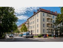 Wohnstraße in einer Stadt mit mehrstöckigen, gelb verputzten Wohngebäuden und roten Ziegeldächern. Am Straßenrand stehen geparkte Autos, Bäume säumen die Straße, und es gibt eine Bushaltestelle mit einem Wartehäuschen.