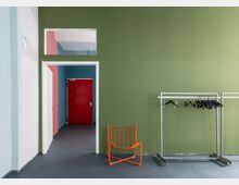 Ein minimalistischer Innenraum mit grüner Wand, einer offenen Tür im weißen Türrahmen, die einen Blick auf eine rote Tür in einem bläulichen Raum freigibt. Rechts steht eine Garderobe mit leeren schwarzen Kleiderbügeln, daneben ein orangefarbener Drahtstuhl.