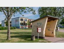 Schulgelände mit einem flachen, modernen Gebäude im Hintergrund, auf dessen Fassade das Wort „Schule“ in großen bunten Buchstaben steht. Im Vordergrund eine überdachte, gelbliche Sitznische mit Graffiti und zwei Süßwarenautomaten sowie Grünflächen und Bäume.