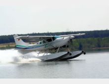 Ein Wasserflugzeug beim Start auf einer ruhigen Wasseroberfläche, umgeben von einem bewaldeten Ufer im Hintergrund. Der Flieger hat Schwimmer statt Räder und erzeugt eine Gischt beim Abheben.