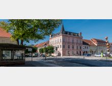 Städtische Straßenszene mit einem historischen, rosafarbenen Gebäude mit Turmspitze im Zentrum, flankiert von weiteren Gebäuden mit roten Ziegeldächern. Im Vordergrund befinden sich eine überdachte Infotafel, Bäume und eine moderne Skulptur aus übereinander gestapelten Kugeln.