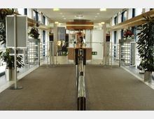 Eingangsbereich eines Gebäudes mit Drehkreuzen, einem Tresen im Hintergrund und dekorativen Pflanzen an den Seiten. Der Raum ist mit Glaswänden und Fluchtwegeschildern ausgestattet.