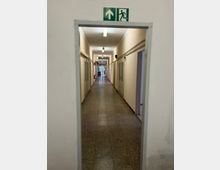 Langer, schmaler Innenflur eines Gebäudes mit Fliesenboden, hellen Wänden und mehreren Türen auf beiden Seiten, beleuchtet durch Deckenleuchten. Am Eingang hängt ein grünes Notausgangsschild, am Ende des Gangs ist eine verglaste Tür zu sehen.