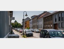 Eine gepflasterte Wohnstraße mit parkenden Autos auf beiden Seiten, umgeben von niedrigen Häusern mit unterschiedlichen Fassadenstilen. Laternen und ein kleiner Gehweg befinden sich auf der linken Seite, und im Hintergrund sind Bäume sichtbar.