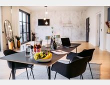 Moderner Besprechungsraum mit einem schwarzen Tisch, darauf Laptops, Notizbücher, Wasserflaschen, Saftflaschen und eine Schale mit Obst. Der Raum ist hell mit Holzboden, großen Fenstern, dekorativen Pflanzen und minimalistischem Design.