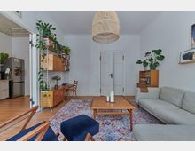 Wohnzimmer mit minimalistischer Einrichtung, Holzmöbeln und Pflanzen. Im Raum stehen ein graues Sofa, ein Holz-Couchtisch, ein bunter Teppich und Regale mit Büchern; eine offene Tür führt zur Küche mit Kühlschrank.