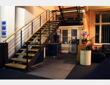 Innenraum eines modernen Bürogebäudes mit einer Treppe aus Holzstufen und Metallgeländer, die nach oben führt. Neben der Treppe befindet sich eine kleine Kaffeestation, eine Sitzgruppe mit schwarzen Ledersesseln und mehrere Pflanzen; im Hintergrund sind Gittertüren und ein großes Fenster sichtbar.