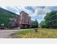 Backstein-Gebäude eines ehemaligen Industriekomplexes mit teilweise von Efeu bewachsenen Fassaden, großen Fenstern und einem kleinen Vorplatz mit Rasen und Bäumen; im Vordergrund eine gepflasterte Straße und eine Laterne.