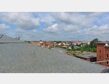 Blick über eine flache Dachfläche mit Kiesbedeckung auf eine Kleinstadt mit roten Ziegeldächern und Gebäuden, teils von Bäumen umgeben. Im Hintergrund sind ein Kirchturm und weitere historische Gebäude sichtbar, unter einem bewölkten Himmel.