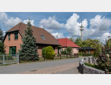 Straßenszene in einem Wohngebiet mit zwei Einfamilienhäusern, eines aus roten Ziegeln und eines in heller Farbe, beide mit roten Satteldächern. Im Vordergrund sind Vorgärten mit Zäunen und Pflanzen, im Hintergrund ein hoher Sendemast und Bäume zu sehen.