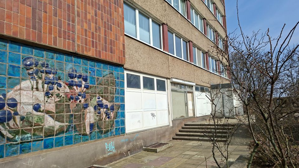 Außenansicht eines mehrstöckigen Wohn- oder Bürogebäudes mit rau verputzter Fassade und langen Fensterreihen. Im Vordergrund ein Eingangsbereich mit Treppenstufen und Gehweg, daneben eine große blaue Fliesenwand mit einem floralen Mosaik sowie kahle Sträucher.