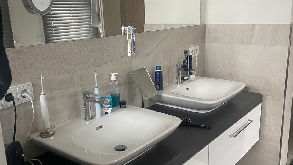 Modernes Badezimmer mit einem Doppelwaschbecken auf einer schwarzen Arbeitsplatte, darunter weiße Schubladen. Im Hintergrund befinden sich ein großer Spiegel, hellgraue Wandfliesen und ein Fenster mit geschlossenen Jalousien.