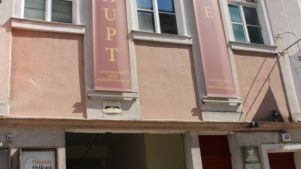 Fassade eines mehrgeschossigen Altbaus in hellem Rosa mit weißen Fensterrahmen und zwei hohen vertikalen Bannern mit der Aufschrift „MÜHLENHAUPT“ und „HÖFE“. Im Erdgeschoss führt eine breite Durchfahrt in einen Innenhof; neben dem Eingang stehen Schautafeln und Beschilderungen.