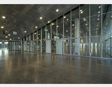 Großer, moderner Innenraum mit glänzendem dunklem Boden, einer Decke aus Metallpaneelen und einer durchgehenden Glaswand mit Türen. Die Glaswand gibt den Blick auf einen ähnlich gestalteten angrenzenden Bereich frei.