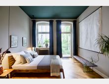 Modernes Schlafzimmer mit Holzboden, einem schlichten Holzbett mit grauen und weißen Textilien sowie einem Nachttisch links. Große Fenster mit dunkelblauen Vorhängen lassen Tageslicht herein, daneben stehen Zimmerpflanzen und eine minimalistische Bank.