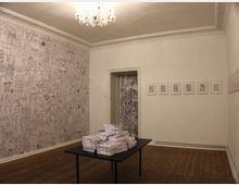 Ein minimalistischer Ausstellungsraum mit Holzboden und hell gestrichenen Wänden. An der Rückwand ein Türdurchgang, umgeben von Kunstwerken, darunter eine Wand mit Zeichnungen, mehrere gerahmte Bilder an der rechten Wand und ein schwarzer Tisch in der Mitte mit gestapelten Büchern.