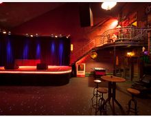 Ein kleiner Veranstaltungsraum mit einer Bühne, deren Hintergrund aus einem dunkelblauen Vorhang besteht und von Scheinwerfern beleuchtet wird. Im Vordergrund stehen Tische und Barhocker, rechts eine Treppe zu einer oberen Galerie, und an der Ziegelwand hängen Retro-Dekorationen; ein Tischkicker befindet sich ebenfalls im Raum.