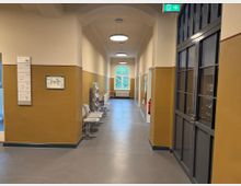 Ein Flur in einem öffentlichen Gebäude mit gelb gestrichenen Wänden und abgerundeter Decke. Auf der rechten Seite befinden sich große Glastüren, links ein Orientierungsschild und ein Lageplan; am Ende des Flurs ist ein großes Fenster mit Blick nach draußen zu sehen.