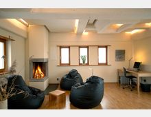 Gemütlicher Raum mit Kamin, mehreren schwarzen Sitzsäcken, einem kleinen Holztisch und hölzernem Schreibtisch samt Computer. Die Wand hat vier Fenster mit Holzrahmen, und die Decke zeigt sichtbare Balken mit integrierter Beleuchtung.