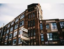 Backstein-Gebäude mit großen Fensterfronten und Industriefassaden, ergänzt durch mehrere Holzwegweiser mit Aufschriften wie „Fabrik 23“, „Office“ und „Werkloft“. Der Himmel ist teilweise bewölkt.