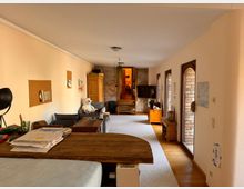 Wohnraum mit hellbeigen Wänden, einer Mischung aus Teppich- und Holzboden und mehreren großen Fenstern mit Rundbögen auf der rechten Seite. Der Raum enthält eine Sofaecke, Holzmöbel, einen Fernseher, Dekorationen an den Wänden und einen erhöhten Bereich im Hintergrund mit Backsteinwänden.