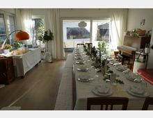 Wohnzimmer mit einem großen Esstisch, der für eine Feier gedeckt ist, mit weißer Tischdecke, gefalteten Servietten, Gläsern und Kerzen. Im Hintergrund sind helle Vorhänge, große Fenster mit Blick nach draußen, ein Klavier und eine Stehlampe sichtbar.