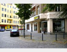 Straßenecke in einer Wohngegend mit gepflastertem Gehweg, umgeben von mehrstöckigen Gebäuden in gelber und grauer Fassade. An der rechten Seite befindet sich ein Geschäft mit großen Schaufenstern, vor dem eine Person steht, sowie parkende Autos entlang der Straße.