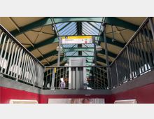 Bahnhofshalle mit einem überdachten Dach aus grünen Metallstreben und verglasten Paneelen, einem Hinweisschild „Dänenstr.“ sowie eisernen Geländern. Im Vordergrund ein Aufzug und eine Treppe, im Hintergrund sicheres und beleuchtetes Bahnsteigdesign.