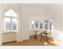 Heller Raum mit einem Erkerfenster aus mehreren vertikalen Sprossenfenstern, einem kleinen runden Holztisch und einem Korbstuhl mit Fellauflage. Links ein spitzbogiges Sprossenfenster mit weißen Vorhängen, der Boden ist mit Teppich ausgelegt.