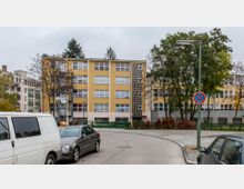 Ein vierstöckiges Wohngebäude mit gelber und weißer Fassade, großen Fenstern und einem vertikal angeordneten Glasbaustein-Element in der Mitte. Im Vordergrund sind geparkte Autos, ein Gehweg, ein Straßenschild mit Parkverbot und vereinzelt Bäume sichtbar.