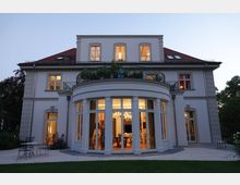 Zweistöckige weiße Villa mit rotem Walmdach und großen Fenstern, darunter ein halbrunder Erker mit verzierten Balkongeländern und großen Glasfronten. Die beleuchteten Innenräume sind sichtbar, umgeben von einem Garten mit Terrasse und Gartenmöbeln.