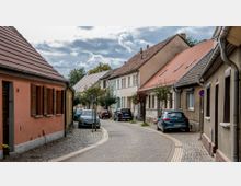 Gepflasterte Straße in einem Wohngebiet mit kleinen, eng aneinander stehenden Häusern mit roten Ziegeldächern. Entlang der Straße sind geparkte Autos und vereinzelt kleine Bäume zu sehen.