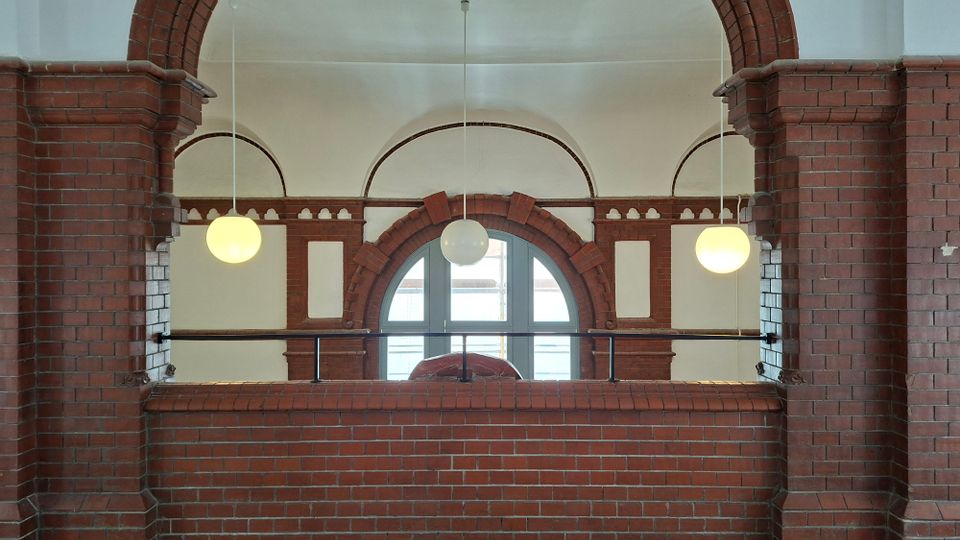 Innenraum mit rotbraunen Backsteinbögen und gemauerten Brüstungen, davor eine Galerie mit schwarzem Geländer. In der Mitte befindet sich ein großes Rundbogenfenster, flankiert von kleineren Bögen, mit drei hängenden Kugelleuchten vor einer hell verputzten Decke.
