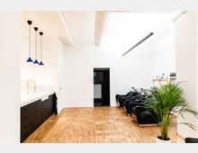 Moderner Friseursalon mit hellem Holzboden, weißen Wänden und schwarzem Mobiliar. Links befindet sich eine Arbeitsfläche mit weißen Fliesen und blauen Hängeleuchten, rechts sind mehrere schwarze Salonwaschplätze sowie Regale mit Produkten zu sehen, im Vordergrund steht eine grüne Zimmerpflanze.