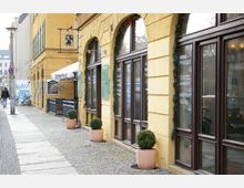 Straßenszene in einer europäischen Stadt mit einem gelben Gebäude und großen Fenstern auf der rechten Seite, dekoriert mit grünen Girlanden. Vor den Fenstern stehen Terrakotta-Blumentöpfe mit runden Buchsbaumsträuchern; links führt ein gepflasterter Bürgersteig mit Fahrrädern, einer Graffiti-Wand und einem Café mit Markise.