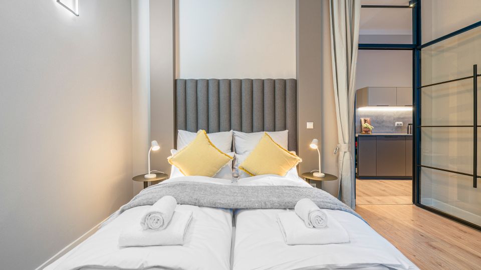 Modernes Schlafzimmer mit Doppelbett, hohem grauem gepolstertem Kopfteil, gelben Kissen und Nachttischen mit Leselampen. Rechts führt eine Glas-Schiebetür mit Vorhang in eine kleine Küche mit dunklen Schränken und Holzfußboden.