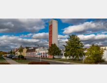 Freistehendes Feuerwehrgebäude mit einem hohen, schmalen Turm in Rot und Weiß, umgeben von Bäumen und geparkten Autos entlang einer Straße. Der Himmel ist bewölkt mit vereinzelten blauen Stellen.