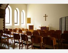 Ein kleiner Andachtsraum mit schlichten Holzbänken, einer Kanzel vorne und einem Holzkreuz an der weißen Wand. Links sind drei schmale, bogenförmige Fenster, und rechts steht ein mit violettem Tuch bedeckter Tisch mit Kerzen und einer Bibel.