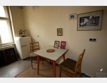 Ein einfacher, altmodischer Küchenraum mit einem kleinen weißen Tisch und drei Holzstühlen. Im Hintergrund steht ein weißer Kühlschrank neben einer Heizung und einem großen Fenster, während an der beige gestrichenen Wand Bilder und Dekorationen hängen.