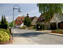 Wohnstraße in einem ruhigen Vorort mit kleineren Einfamilienhäusern, Vorgärten und gepflastertem Gehweg, umgeben von Bäumen und Sträuchern; im Hintergrund ein größeres Gebäude mit rotem Ziegeldach und einem kleinen Turm.