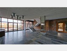 Großzügige, moderne Eingangshalle mit grauem Steinboden, Backsteinwänden und einer zentralen Treppe mit Edelstahlgeländer. Links sorgen bodentiefe Glasfronten für Tageslicht, rechts führen Glastüren in angrenzende Räume; an der Decke hängen runde Leuchten.