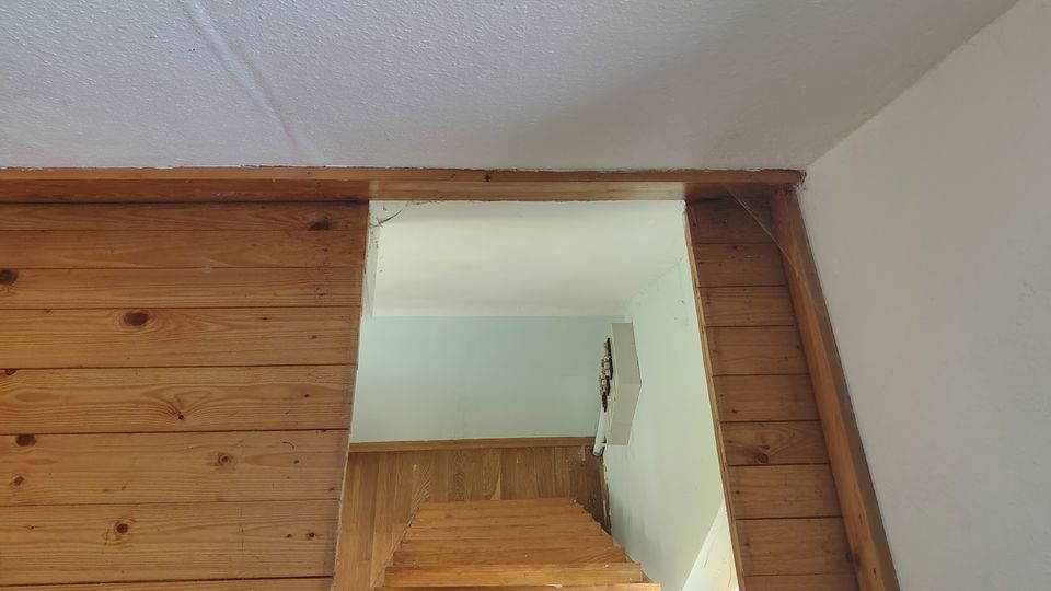 Blick von unten auf eine schmale Holztreppe, die durch einen rechteckigen Durchgang nach oben führt. Die Wände und Verkleidungen sind aus Holzpaneelen, darüber eine weiße, strukturierte Decke und angrenzende weiße Wandflächen.